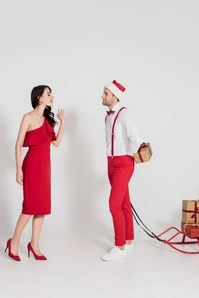 Brunette woman in red dress standing near man in santa hat hiding present and sleigh on grey background — Stock Photo
