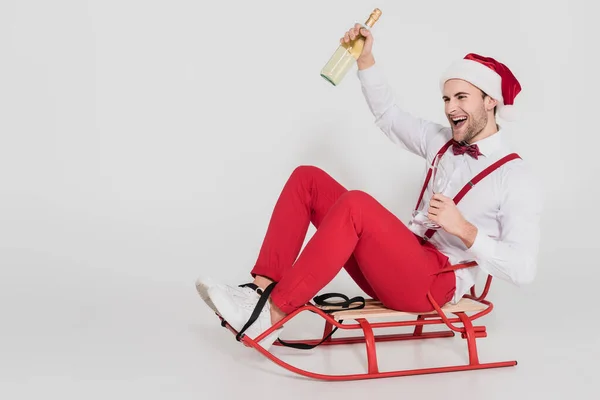 Cheerful man in santa hat holding glass and bottle of champagne on sleigh on grey background — Stock Photo