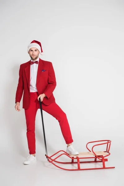 Young man in red suit and santa hat holding sleigh on grey background — Stock Photo