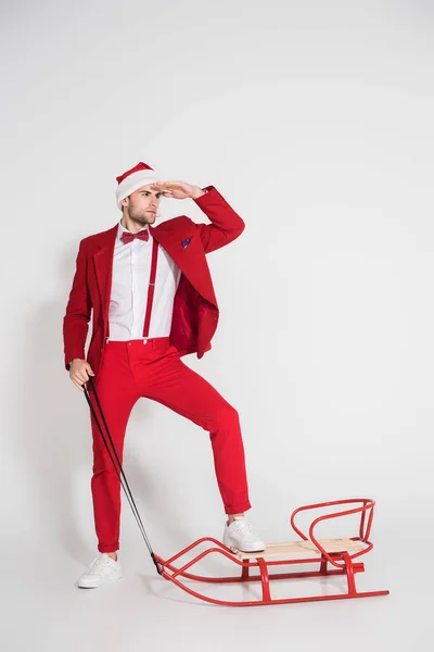 Man in red suit and santa hat looking away near sleigh on grey background — Stock Photo