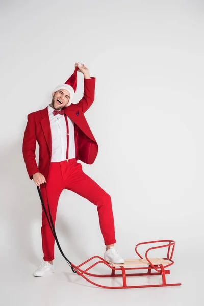 Homme excité en costume rouge tenant un chapeau de Père Noël près du traîneau sur fond gris — Photo de stock
