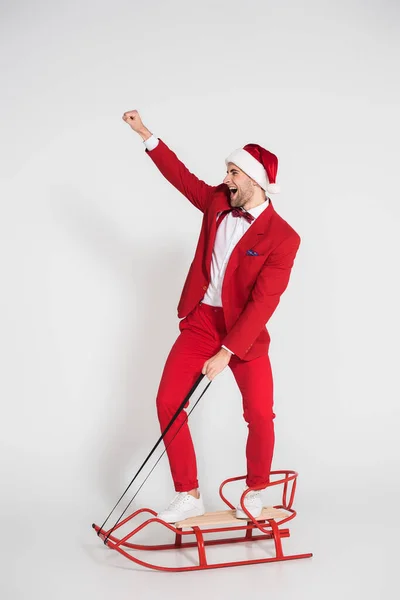 Cheerful man in suit and santa hat standing on sleigh on grey background — Stock Photo