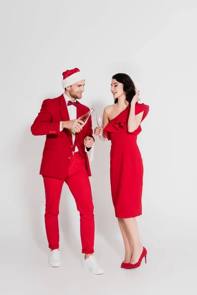 Homem sorridente de terno e chapéu de Papai Noel segurando garrafa de champanhe perto de mulher com vidro no fundo cinza — Fotografia de Stock