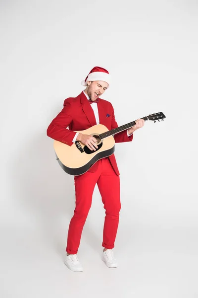 Aufgeregter Mann im Anzug und roter Weihnachtsmütze spielt Akustikgitarre auf grauem Hintergrund — Stockfoto