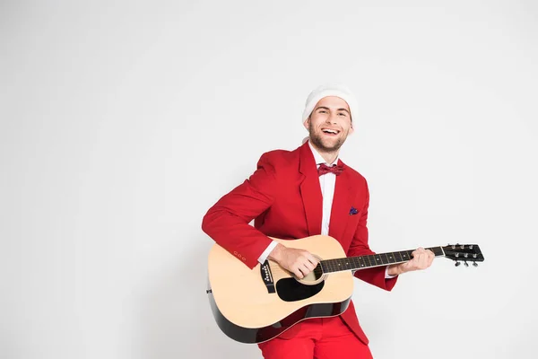 Jeune homme en costume et chapeau de Père Noël souriant à la caméra tout en jouant de la guitare acoustique sur fond gris — Photo de stock