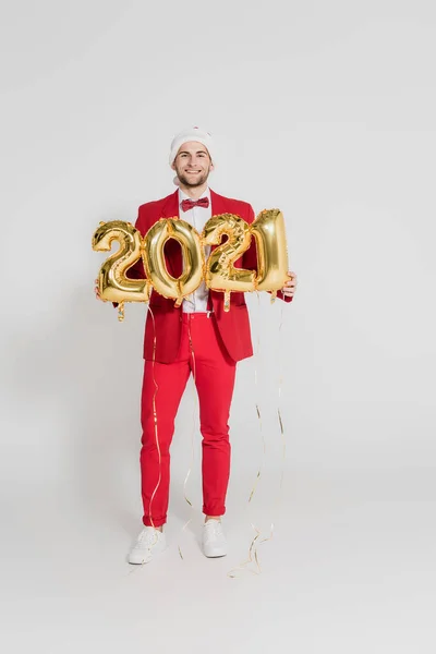 Homme souriant au chapeau de Père Noël tenant des ballons en forme de chiffres 2021 sur fond gris — Photo de stock