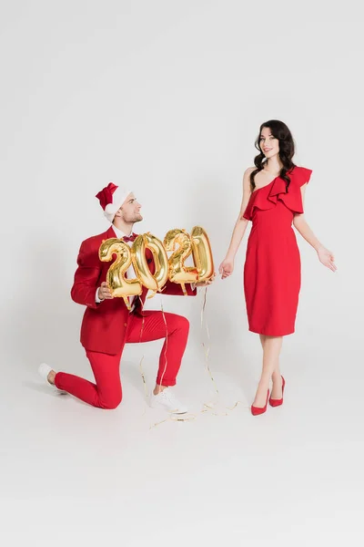 Fröhlicher Mann mit Weihnachtsmütze steht auf einem Knie und hält Luftballons in Form von Ziffern neben einer Frau auf grauem Hintergrund — Stockfoto