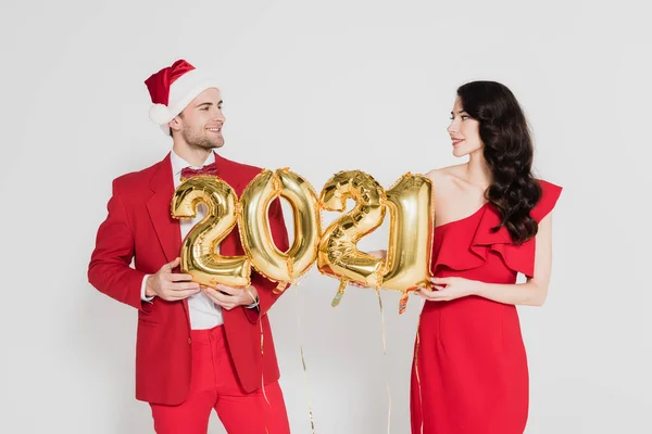 Hombre sonriente en sombrero de santa y mujer en vestido rojo sosteniendo globos en forma de números 2021 aislados en gris - foto de stock