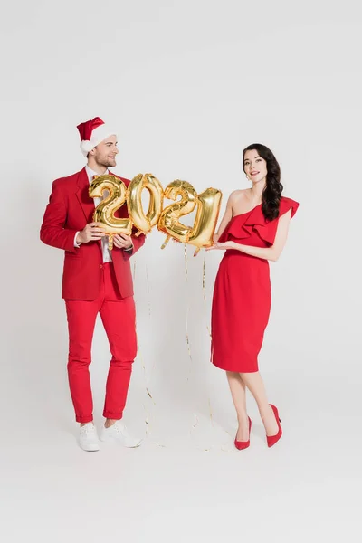 Mujer morena en vestido rojo sosteniendo globos en forma de 2021 números cerca de hombre en sombrero de santa sobre fondo gris - foto de stock