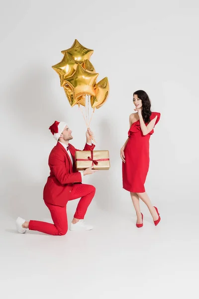 Smiling man in santa hat holding balloons and present near woman in red dress on grey background — Stock Photo