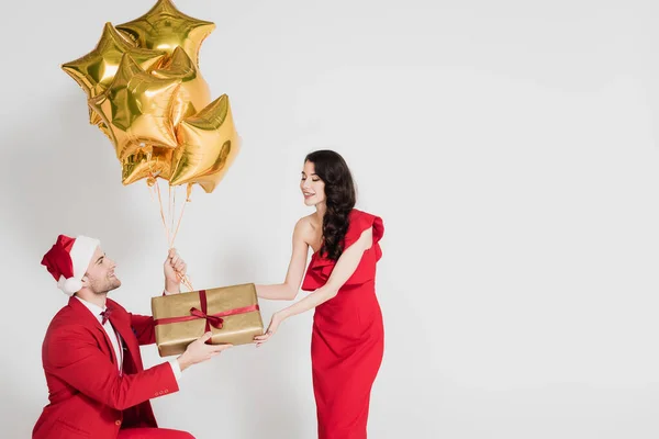 Homme au chapeau santa tenant des ballons dorés et cadeau près d'une femme souriante sur fond gris — Photo de stock