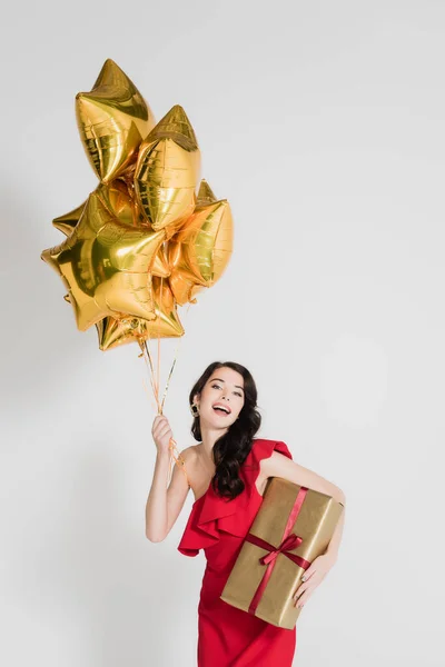 Mujer alegre en vestido rojo sosteniendo presente y globos dorados sobre fondo gris - foto de stock
