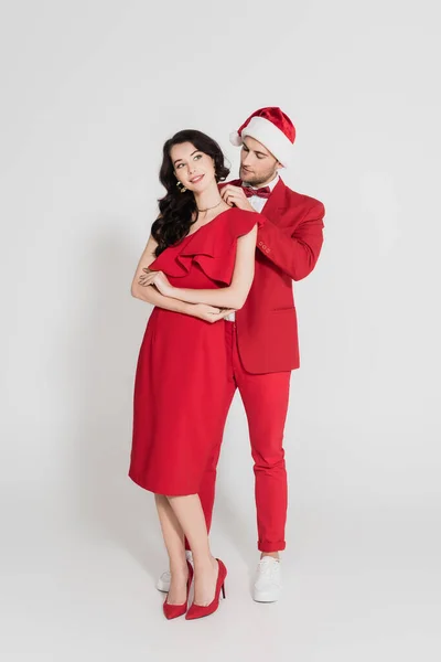 Homme en costume et chapeau de Père Noël portant un collier sur une femme brune souriante sur fond gris — Photo de stock