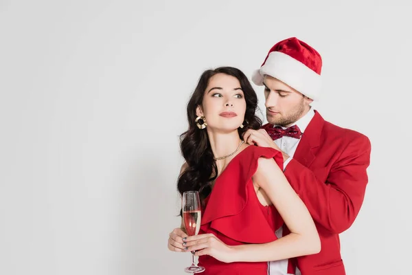 Mann mit Weihnachtsmütze und Fliege trägt Halskette auf Frau im Kleid mit Glas Champagner auf grauem Hintergrund — Stockfoto