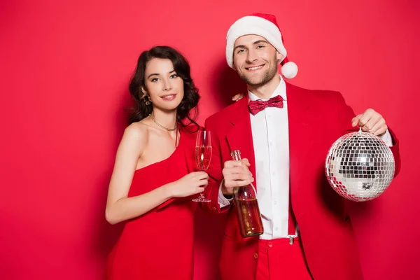 Усміхнена пара з диско-кулькою, шампанським та капелюхом Санти дивиться на камеру на червоному тлі — стокове фото