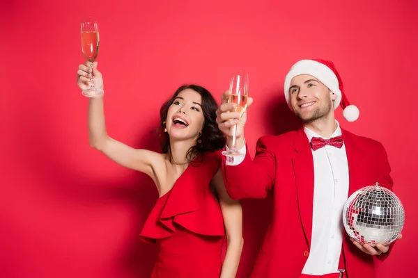 Cheerful couple with disco ball and santa hat holding glasses of champagne on red background — Stock Photo