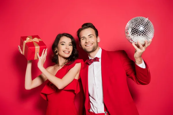 Sonriente pareja con regalo y bola disco mirando a la cámara sobre fondo rojo - foto de stock