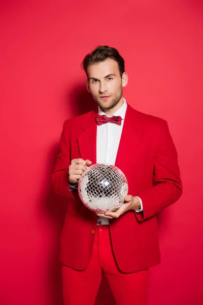 Junger Mann im Anzug hält Discokugel in der Hand und blickt in Kamera auf rotem Hintergrund — Stockfoto
