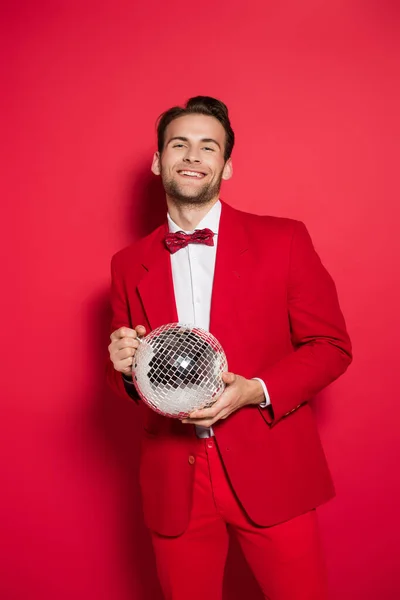 Hombre sonriente en traje rojo sosteniendo bola disco sobre fondo rojo - foto de stock