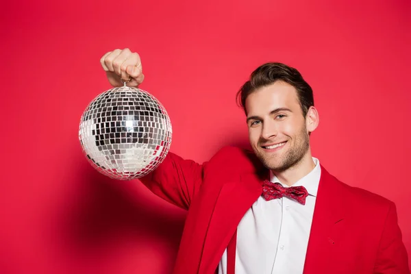 Homme élégant en costume rouge souriant tout en tenant la balle disco sur fond rouge — Photo de stock