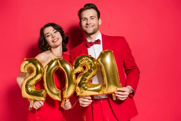 Alegre pareja sosteniendo globos en forma de 2021 números sobre fondo rojo - foto de stock