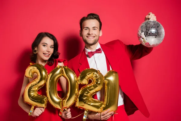 Cheerful young couple holding disco ball and balloons in shape of 2021 numbers on red background — Stock Photo