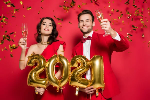 Coppia sorridente con bicchieri di champagne che tiene palloncini a forma di 2021 numeri sotto i coriandoli che cadono su sfondo rosso — Foto stock