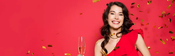 Smiling woman holding glass of champagne under falling confetti on red background, banner — Stock Photo