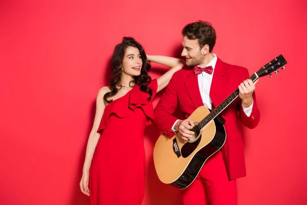 Mulher sorridente de vestido olhando para o namorado tocando guitarra acústica no fundo vermelho — Fotografia de Stock