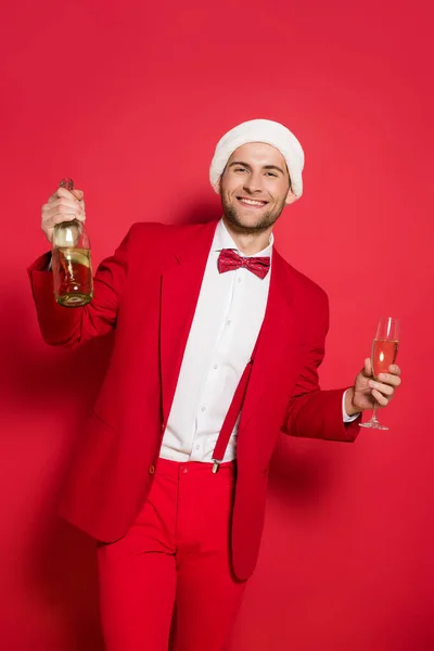 Elegante hombre en sombrero de santa celebración de champán y sonriendo a la cámara sobre fondo rojo - foto de stock
