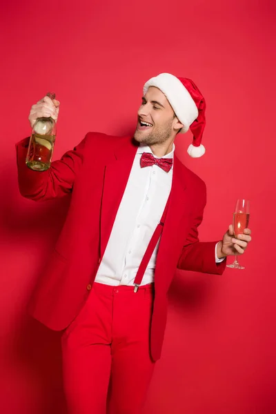 Young man in santa hat and red suit holding bottle and glass of champagne on red background — Stock Photo
