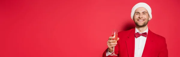 Mann mit Weihnachtsmütze und Anzug lächelt in die Kamera, während er ein Glas Champagner auf rotem Hintergrund hält, Banner — Stockfoto