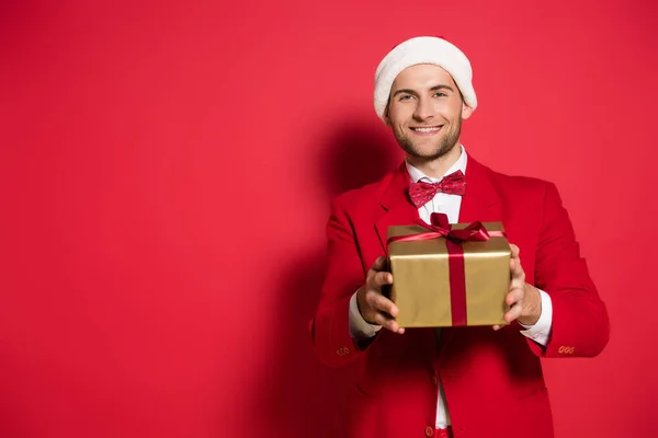 Positive man in santa hat and suit holding present with bow on red background — Stock Photo
