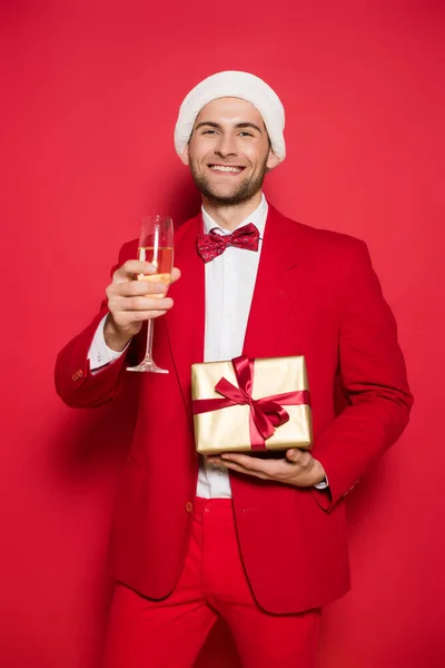 Homme souriant au chapeau de Père Noël tenant présent et verre de champagne sur fond rouge — Photo de stock