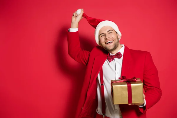 Homem alegre segurando chapéu de Papai Noel e presente no fundo vermelho — Fotografia de Stock