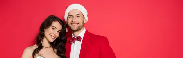 Усміхнена жінка дивиться на камеру біля хлопця в капелюсі Санта ізольовано на червоному, банер — стокове фото