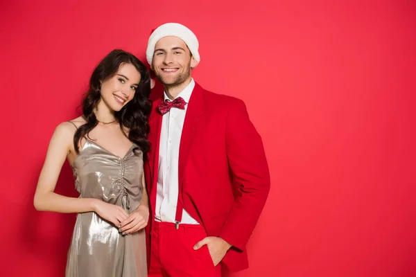 Lächelnde Frau im Kleid, die in die Kamera schaut, neben stilvollem Freund mit Weihnachtsmütze auf rotem Hintergrund — Stockfoto