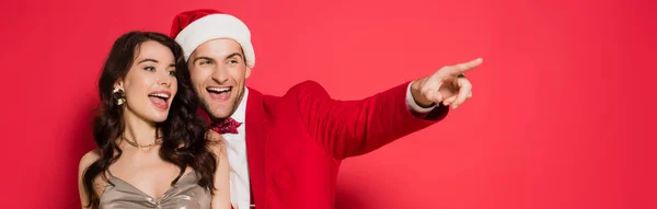 Cheerful woman standing near boyfriend in santa hat pointing with finger on red background, banner — Stock Photo