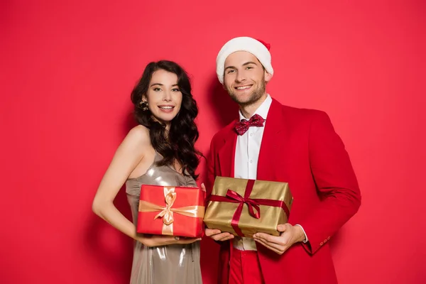 Couple joyeux avec santa chapeau tenant des boîtes-cadeaux sur fond rouge — Photo de stock