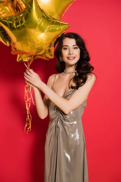Brünette Frau im Kleid lächelt in die Kamera, während sie Luftballons auf rotem Hintergrund hält — Stockfoto