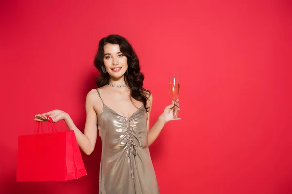 Femme souriante tenant des sacs à provisions et une coupe de champagne sur fond rouge — Photo de stock
