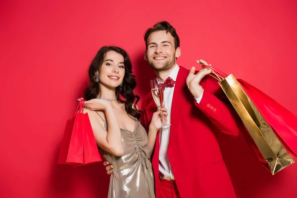 Homem alegre em terno segurando sacos de compras perto da namorada com copo de champanhe no fundo vermelho — Fotografia de Stock