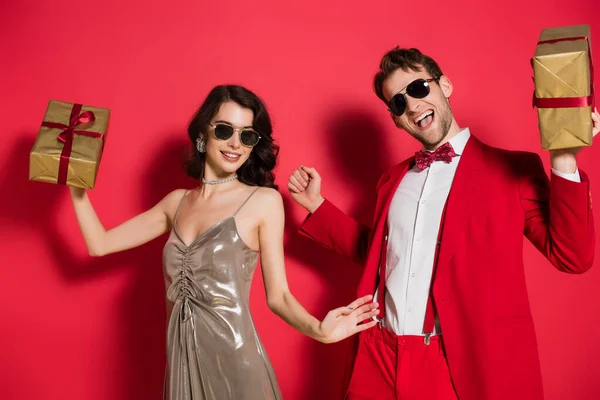 Couple élégant dans des lunettes de soleil tenant des boîtes-cadeaux sur fond rouge — Photo de stock