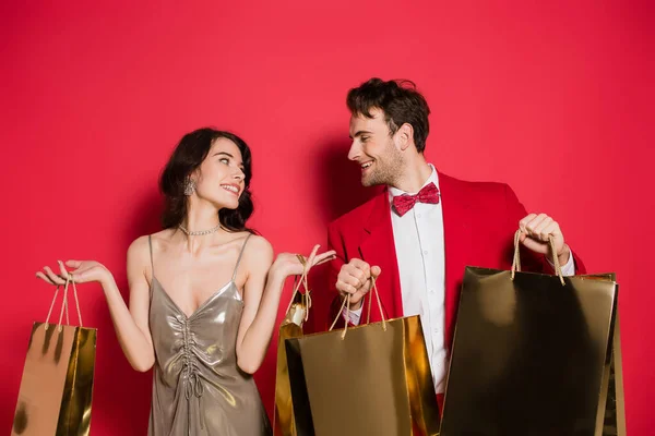 Sorrindo casal olhando um para o outro enquanto segurando sacos de compras no fundo vermelho — Fotografia de Stock