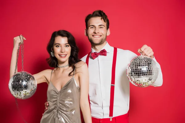 Homme souriant embrassant petite amie tout en tenant des boules de disco sur fond rouge — Photo de stock