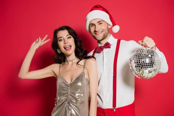 Mujer alegre mostrando signo de paz cerca de novio en sombrero de santa con bola disco sobre fondo rojo - foto de stock