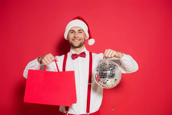 Smiling man in santa hat holding disco ball and shopping bags on red background — Stock Photo