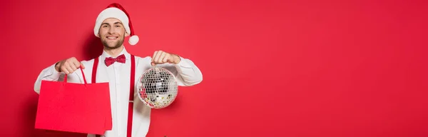 Positive man in santa hat holding disco ball and shopping bags on red background, banner — Stock Photo