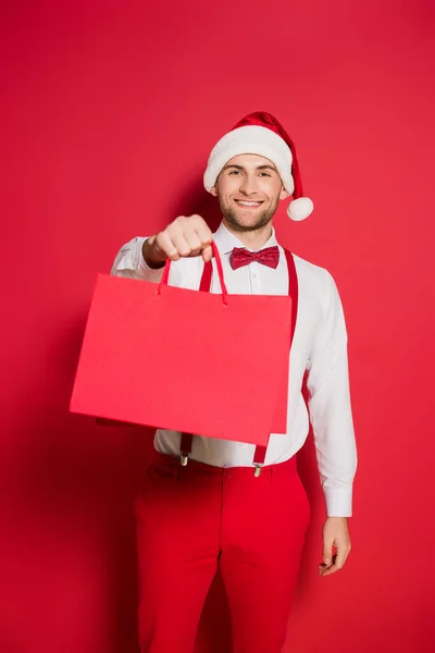 Stylischer Mann mit Weihnachtsmütze lächelt, während er Einkaufstüten auf rotem Hintergrund hält — Stockfoto