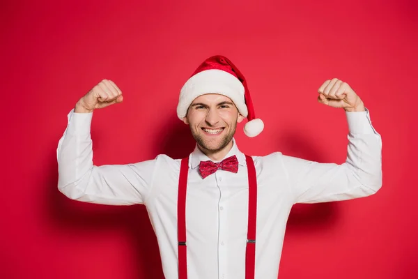 Stylish man in santa hat showing muscles and smiling on red background — Stock Photo